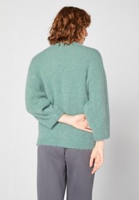 Un pull en maille léger et flou de couleur vert clair, avec une coupe décontractée, des manches trois-quarts et un col haut. Porté avec un pantalon gris.