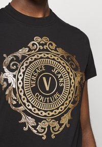 Musta puuvillapaita, jossa on suuri kultafoliojälki, koristeellisia kuvioita, keskeinen logo ja teksti "Versace Jeans Couture."