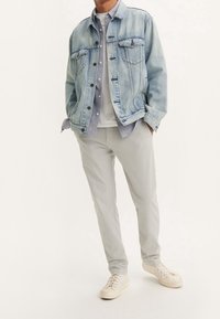 Helles blauer Denimjacke mit zwei Brusttaschen, kombiniert mit einem hellgrauen Hemd und einem weißen T-Shirt; dazu hellgraue Hose und weiße Sneakers.
