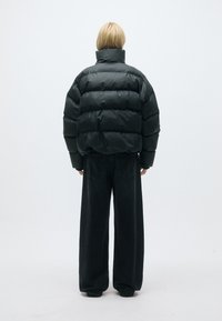 Sort pufferjakke med høj krave og quiltet design; båret med løstsiddende sorte jeans på en ensfarvet baggrund.