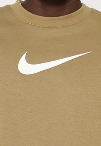 Beige sweatshirt med en vit Nike swoosh-logotyp. Tillverkad av mjuk bomullsblandning med rund hals och ribbade muddar.