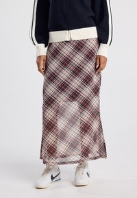 Langer karierten Rock in Beige-, Rot- und Dunkelblau-Tönen, mit einem fließenden Design. Kombiniert mit weißen Sneakers und einem navy-blauen Reißverschlusspullover.