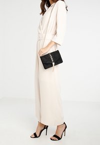 Borsa clutch in suede nero con tracolla a catena argentata e dettaglio con nappina, abbinata a una tuta beige chiaro a gamba larga e tacchi alti neri.