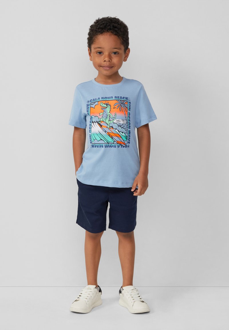 Lichtblauw t-shirt met een kleurrijke strandafbeelding van een krokodil op een surfplank. Gecombineerd met marineblauwe shorts en witte sneakers.