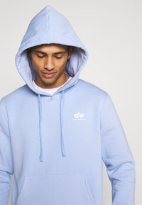 Felpa azzurra chiara in morbido tessuto. Presenta una tasca frontale, cordini e un logo bianco sul petto. Il cappuccio è foderato con una tonalità più chiara.
