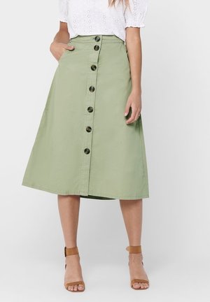Woman wearing a knee-length light green skirt with front buttons, a white eyelet blouse, and tan ankle-strap sandals, standing with one hand in pocket.