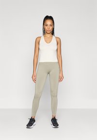 Λευκό αθλητικό αμάνικο μπλουζάκι συνδυασμένο με ελαιόχρωμα κολάν. Το μπλουζάκι έχει εφαρμοστή σχεδίαση, ενώ τα κολάν διαθέτουν λεία υφή και ελαφριά γυαλάδα.