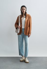 Blazer marrón de cuero sintético sobre una blusa de punto blanca, jeans de pierna ancha azules con dobladillo deshilachado y zapatos beige gruesos con acentos florales.