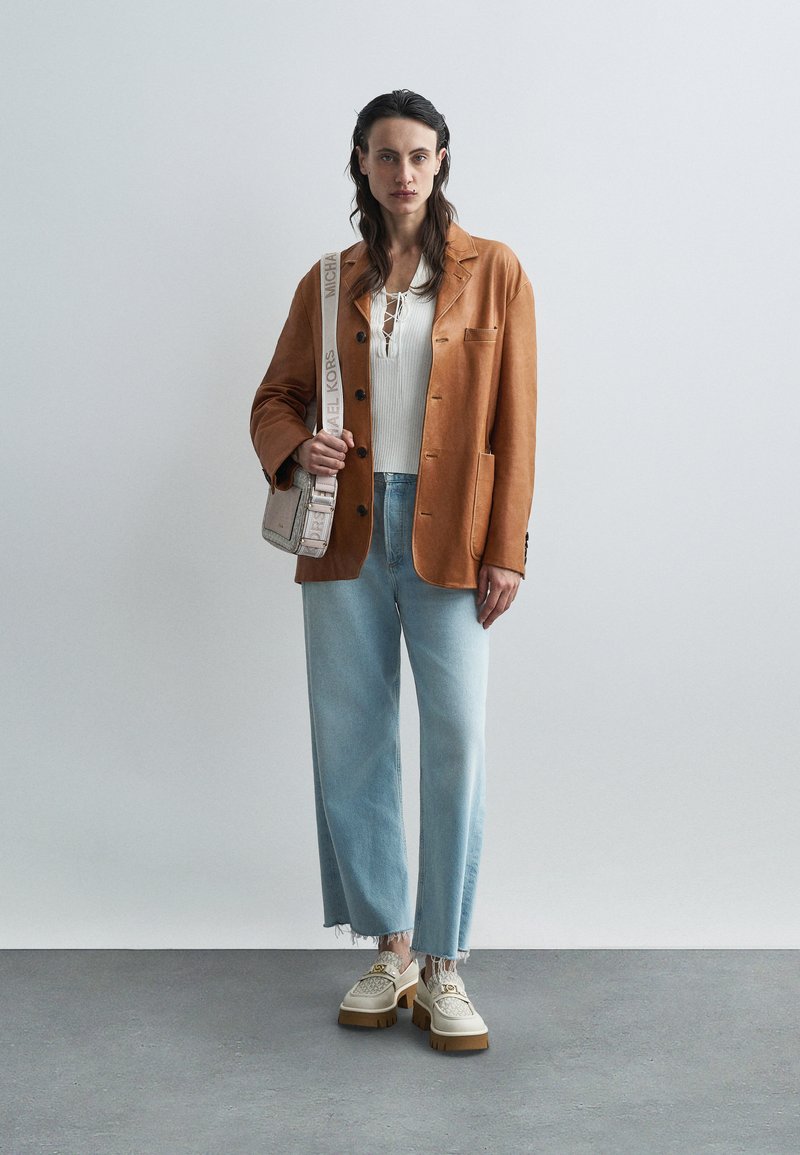 Blazer marrón de cuero sintético sobre una blusa de punto blanca, jeans de pierna ancha azules con dobladillo deshilachado y zapatos beige gruesos con acentos florales.