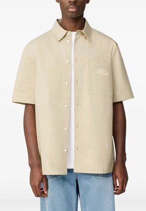 Uomo che indossa una camicia beige a maniche corte con bottoni sopra una T-shirt bianca e jeans azzurri, in piedi davanti a uno sfondo bianco semplice.