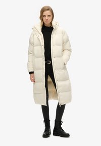 Lang, puffer frakke i off-white, med hætte, trykknapper og en mellemlang længde. Båret over en sort sweater og sorte skinny jeans.