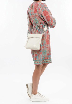 Femme portant une robe colorée à fleurs d'une longueur jusqu'aux genoux, des baskets blanches et un sac bandoulière beige, debout devant un fond blanc uni.