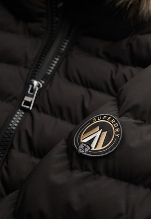 Giacca nera imbottita con una cerniera prominente e una patch circolare con il marchio "Superdry" in oro e bianco.