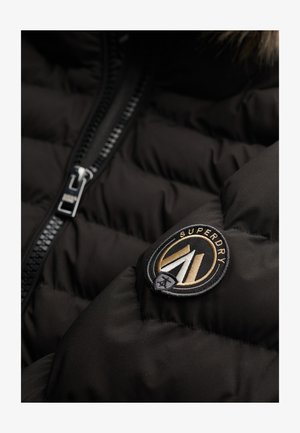 Giacca nera imbottita con una cerniera prominente e una patch circolare con il marchio "Superdry" in oro e bianco.