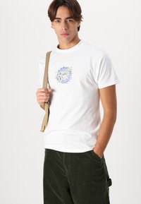 Witte t-shirt met korte mouwen en een blauw grafisch ontwerp op de borst, gecombineerd met groene corduroy broek en een beige schoudertas.