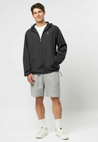 Mees seisab, kandes musta lukuga kapuutsiga jopet, valget T-särki, halli lühikest pükse, valgeid meessokke ja valgeid tossusid, ühevärvilise tausta ees.