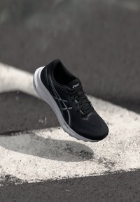 Sorte joggesko med en teksturert overdel, hvit mellomsåle og reflekterende sideakenter. Har lisser og perforeringer for pusteevne.