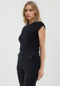 Schwarzes, tailliertes T-Shirt mit kurzen Ärmeln, glatter Textur und Rundhalsausschnitt. Kombiniert mit hoch taillierten schwarzen Hosen mit frontalen Taschen und Knopfverschluss.