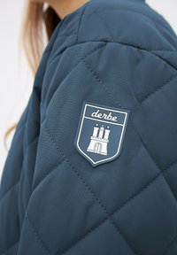 Manche d'une veste matelassée bleu foncé avec un patch logo "derbe" représentant un château blanc à trois tours et des étoiles sur le bras extérieur.