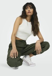 Witte aansluitende mouwloze top gecombineerd met olijfgroene baggybroek en witte hoge sneakers. De outfit heeft een strikkoord in de taille.