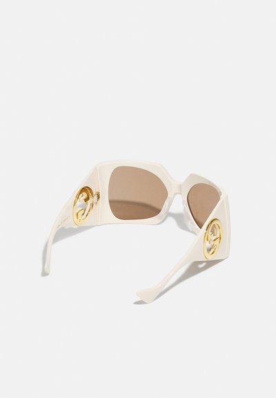 Gafas de sol beige de gran tamaño con lentes marrones. Presentan un distintivo acento dorado en las patillas. Montura de plástico suave con diseño geométrico.