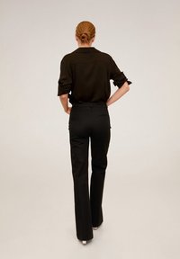 Kobieta z włosami w kok, ubrana w czarną bluzkę z długim rękawem z podwiniętymi rękawami oraz czarne spodnie o wysokim stanie, stojąca z rękami na biodrach, plecami do aparatu.