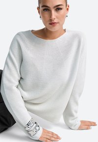 Light gray, loose-fit sweater with a boat neckline. Textured fabric, long sleeves, and silver bangle bracelets accentuate the wrist.