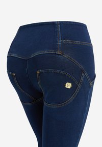 Jeans in denim blu scuro con tasche posteriori curve, cuciture gialle e un piccolo logo sul taschino sinistro.