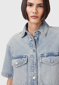 Ljusblå denimskjorta med korta ärmar, knäppning framtill, två bröstfickor och dekorativa nitar längs den centrala knappslån.