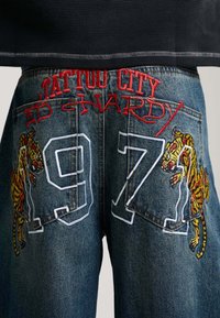 Denim jeans with red "Tattoo City Ed Hardy" embroidery, large white "97" stitching, and embroidered tigers on back pockets.