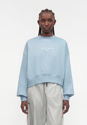 Άτομο με πλεγμένα μαλλιά φοράει ένα ανοιχτό μπλε φούτερ Nike Shox και φαρδιά ανοιχτό γκρι παντελόνια με μικρό μαύρο λογότυπο Nike.