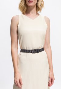 Femeie purtând o rochie crem fără mâneci, cu o centură cu imprimeu leopard gri închis și o cataramă rotundă argintie în talie.