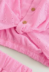 Tissu rose texturé avec des motifs œillets brodés, des boutons en bois et un détail de taille élastique sur un vêtement.