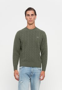Mannlig modell iført olivengrønn kabelstrikket genser med Lacoste-logo og lyseblå slitte jeans, står mot en enkel hvit bakgrunn.