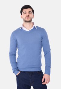 Maglione azzurro chiaro a V, realizzato in tessuto lavorato a maglia, con maniche lunghe e polsini a costine, indossato sopra una camicia bianca con colletto.