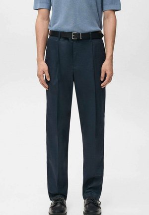 Homme portant un pantalon plissé bleu marine avec ceinture noire, chaussures noires et chemise tricotée à manches courtes bleue, debout devant un fond uni.