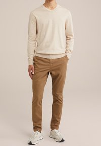 Béžový svetr s výstřihem do V, vyrobený z lehké strukturované látky, v kombinaci s hnědými chinos kalhotami a bílými teniskami, stojící na neutrálním pozadí.