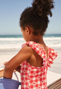 Rot-weiß karierten Badeanzug mit Herzmuster, mit Rüschenträgern, rückenfreiem Design, gehalten mit einem Eimer in der Nähe des Strandes.