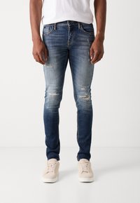 Modré jeans s úzkym strihom, s opotrebovanými oblasťami a svetlým vyblednutím. Skombinované s béžovými teniskami a bielym tričkom.