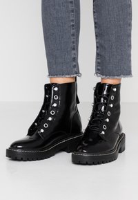 Černé lakované kotníkové boty s stříbrnými očky, silnými černými podrážkami a šněrovacím designem. Kombinované s roztrhanými šedými džíny.