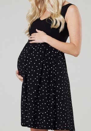 Kobieta w ciąży z długimi blond włosami, ubrana w czarną sukienkę bez rękawów w białe grochy, trzymająca brzuch obiema rękami.