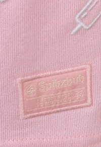 Rosa gestricktes Etikett mit weißem, gesticktem Text "Spitzbub" und Pflegehinweisen, mit einer glatten Textur und klarer Schriftart.
