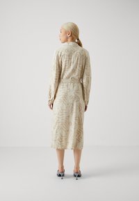 Robe longue beige avec des motifs abstraits, col boutonné, taille ceinturée et manches longues. Portée avec des chaussures à talons gris. Texture lisse.