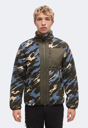 Jeune homme portant une veste polaire à col montant zippée avec un motif camouflage bleu, vert et beige et une poche poitrine zippée verte unie.