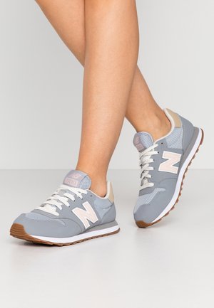 Nogi w jasnoszarych i beżowych butach sportowych New Balance 500 z białymi sznurówkami, stojące na jasnoszarej podłodze na tle jednolitego tła.