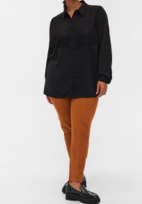 Chemise noire à boutons avec des franges, associée à un pantalon en velours côtelé orange et des mocassins à plateforme noirs. Fond uni.