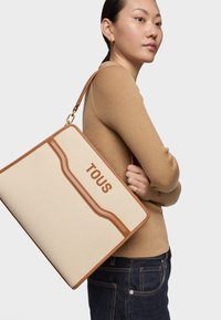 Bolsa de lona con detalles en cuero marrón, logo "TOUS" en relieve, forma rectangular, correa para el hombro y superficie de tela texturizada.