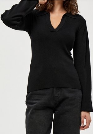 Femme portant un pull noir à manches longues en V avec un col et un pantalon foncé, posant avec un bras levé.