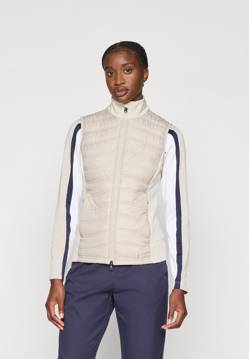 Lett, beige vest med quiltet tekstur, høy krage og glidelås. Har lange hvite ermer med marineblå sidestreker.