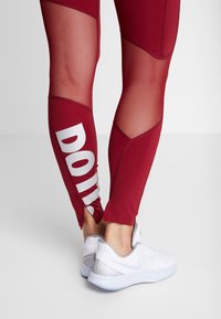 Leggings sportivi rossi con pannelli in rete e testo bianco "FALLO". Abbinati a scarpe da ginnastica bianche con accenti argentati e suola blu chiaro.